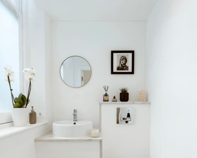 The image shows a small, modern bathroom with a round mirror, a sink, a toilet, and minimal decorative items on the walls and counter.