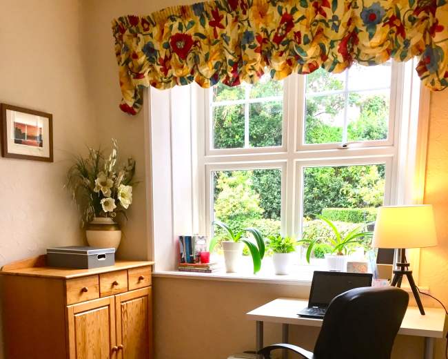 A bright and cozy home office features a desk with a laptop, a chair, a wooden cabinet, and a window adorned with floral curtains, surrounded by potted plants.