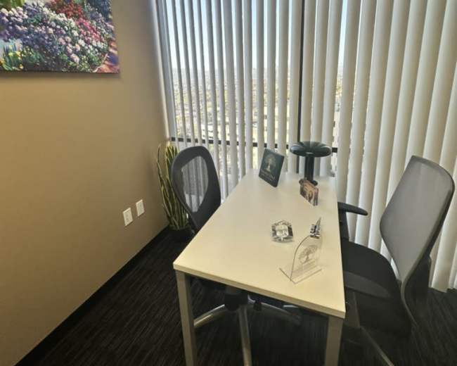 The image shows a small office with a desk and two chairs, featuring a landscape painting on the wall and large windows with vertical blinds.