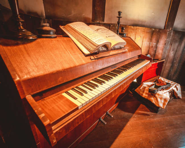 The image shows a wooden piano with sheet music open on top, positioned in a rustic room with wooden walls and a few items nearby.