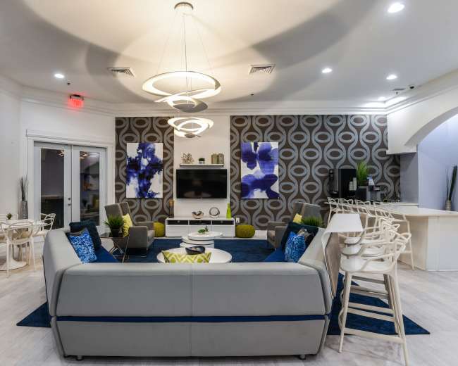 A modern lounge area featuring a gray sectional couch, round coffee tables, and two large artworks on the wall, with a dining space visible in the background.