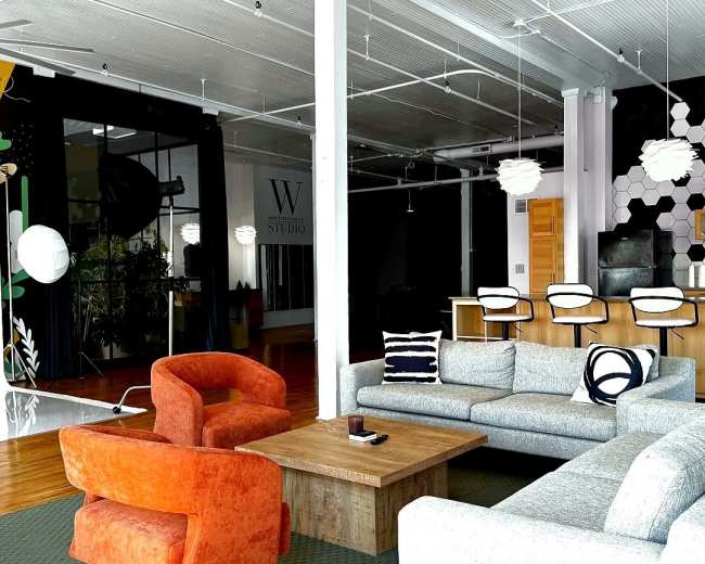 The image shows a modern studio space featuring a mix of seating options, a large wooden table, and a kitchen area with bar stools, all set against a backdrop of contrasting black and vibrant wall designs.