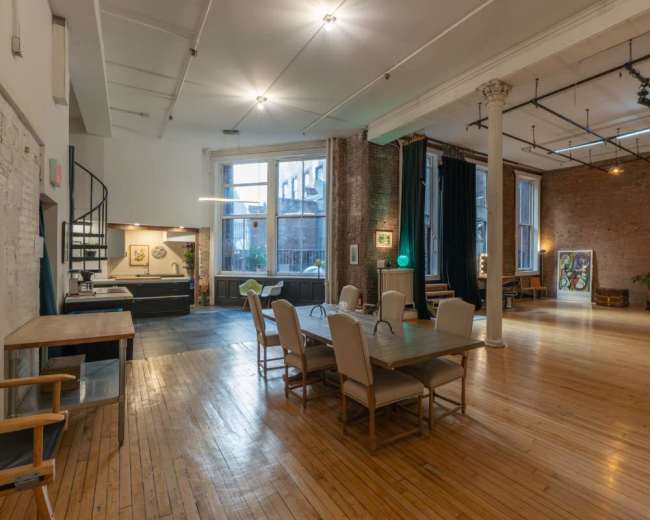 The image shows a spacious, lightly furnished interior with wooden floors, exposed brick walls, a dining table with chairs, and a seating area containing a couch.