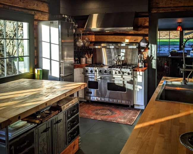 The image shows a modern kitchen with stainless steel appliances, a wooden island, and large windows allowing natural light to enter.