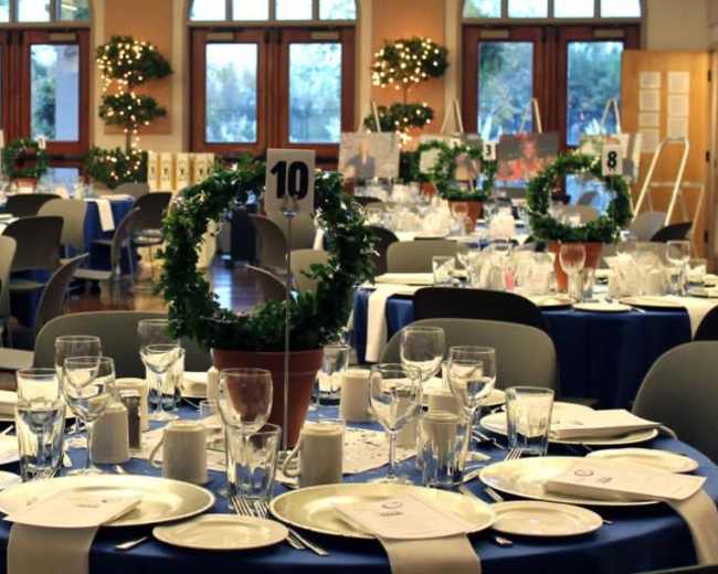 A banquet hall arranged with round tables set for dining, each featuring blue tablecloths, plates, glassware, and decorative centerpieces.