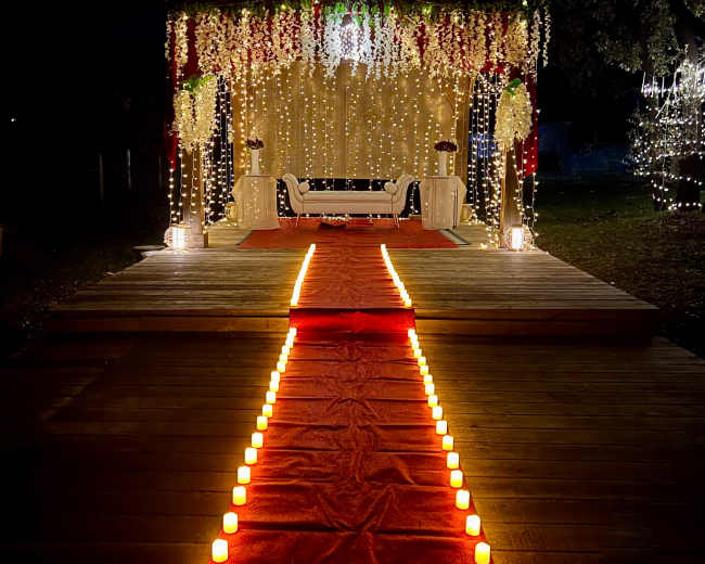 A decorated outdoor stage with a red carpet lined by candles and surrounded by string lights and floral arrangements.
