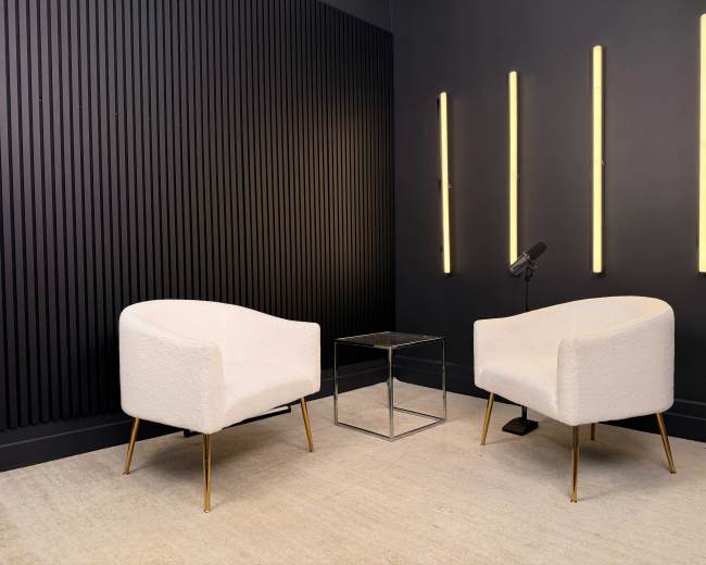 The image depicts a minimalist interior featuring two white armchairs and a glass side table against a dark wall with vertical paneling and illuminated vertical light fixtures.