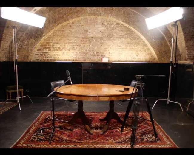 The image shows a dimly lit studio with a wooden table and two chairs, flanked by two studio lights and cameras.