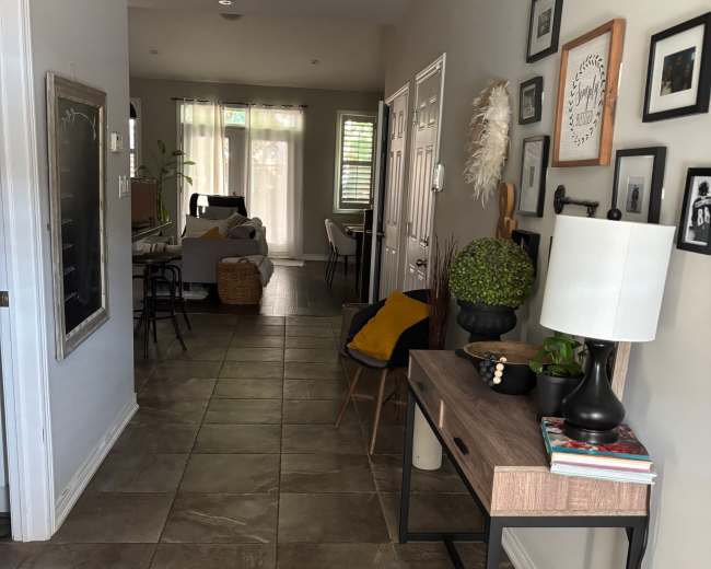 The image shows a hallway with tile flooring, leading into a well-lit living space, and features a console table, decorative items, and framed pictures on the walls.