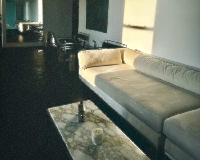A sunlit living space features a light-colored sofa and a marble coffee table, with reflection visible in a mirror on the wall.