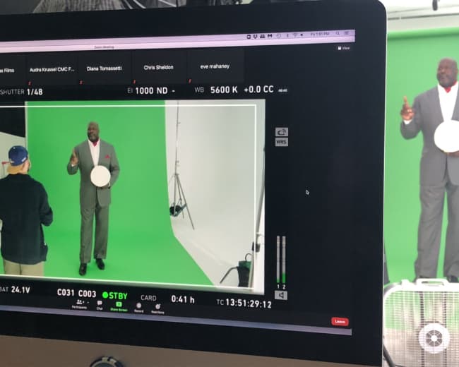 A man in a gray suit stands in front of a green screen while holding a circular object, as he addresses a person off-camera.