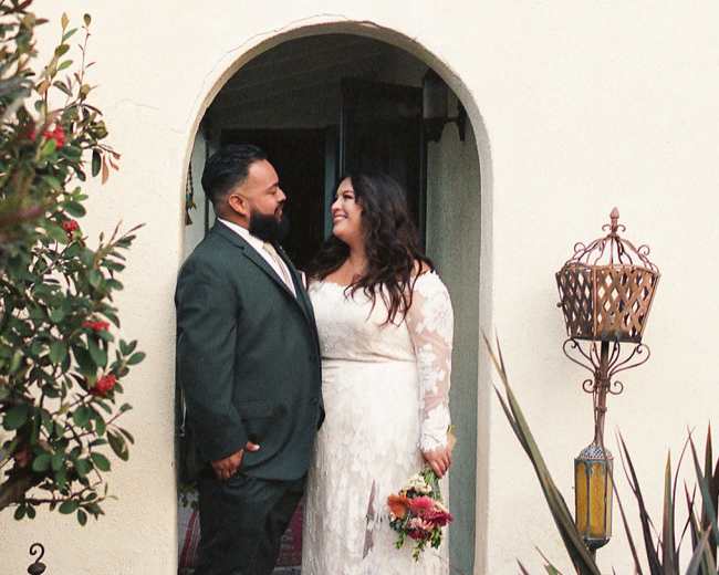 A couple stands in an arched doorway, smiling at each other, while holding a bouquet of flowers.