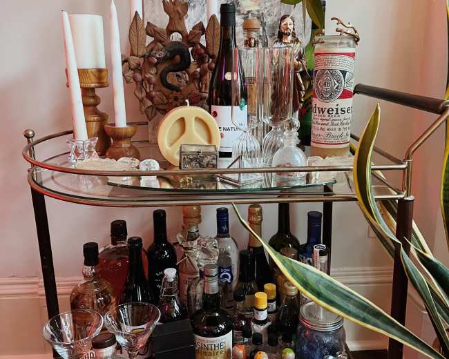 A bar cart displays an assortment of spirits, glassware, decorative candles, and artwork, with a plant nearby.