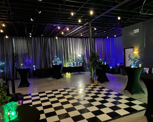 A spacious event venue features a black and white checkered floor, tables set with colorful centerpieces, and soft lighting from overhead fixtures.