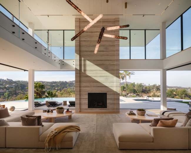 The spacious living room features a tall stone fireplace, large windows overlooking a hillside, and modern furniture arranged around a central area rug.