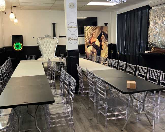 The image shows a spacious event room set up with multiple rows of tables and clear acrylic chairs, featuring decorative lighting and large windows.