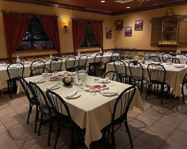 The image shows a dining area with several tables set for a meal, featuring white tablecloths and black chairs, under warm lighting.