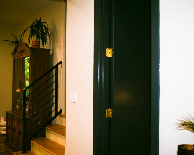 The image shows an interior space featuring a staircase with wooden steps, a green plant near a wall, and a closed black door.