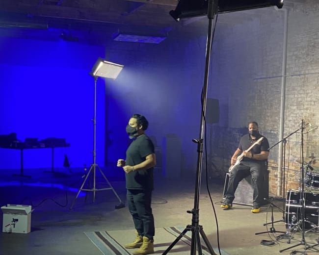 A band performs in a dimly lit warehouse with a blue backdrop and studio lighting.
