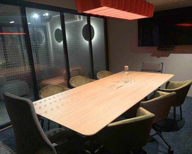 A modern conference room features a large wooden table surrounded by six chairs, with a TV mounted on the wall and glass partitions.