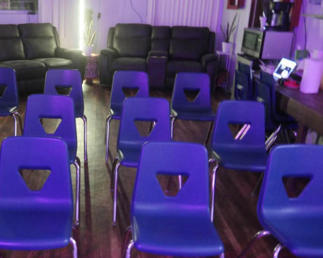 The image shows a room with two dark leather couches and several blue chairs arranged in rows on a wooden floor.
