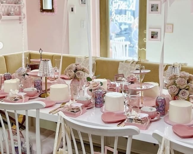 A table is elegantly set with pink decorations, including balloons, flowers, and fine dining ware, in a bright room featuring pastel walls and a cozy layout.