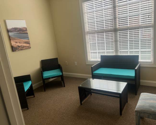 The image shows a small living area with two chairs and a sofa, all featuring turquoise cushions, accompanied by a glass coffee table, and a large window with blinds letting in natural light.