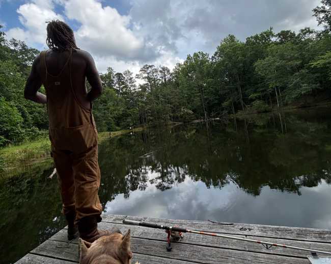 A person in overalls stands on a wooden dock overlooking a calm pond, while a dog rests nearby.