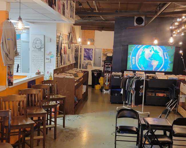 The image shows a record shop interior featuring shelves with vinyl records, display areas for T-shirts, and seating arranged for customers.