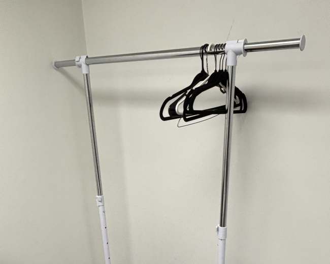A portable clothing rack with metal and plastic components stands against a wall, holding several black hangers.