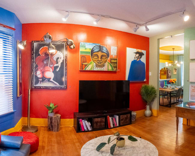 The image shows a vibrant living room with colorful walls, a large television, and various framed artworks, leading to a dining area in the background.