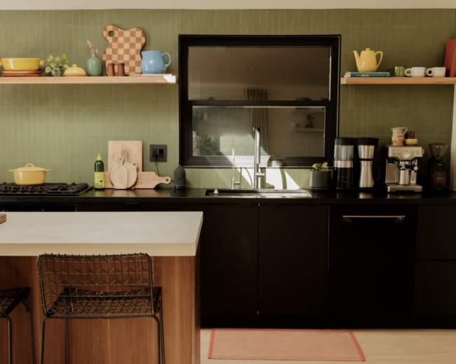 A modern kitchen features a black countertop, wooden bar stools, and a variety of colorful kitchenware displayed on shelves.