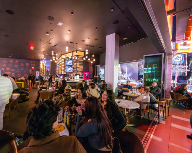The image shows a bustling restaurant interior filled with people seated at tables, illuminated by ambient lighting and decorative fixtures.