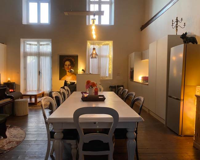 The image shows a spacious, well-lit dining area featuring a long white table, modern kitchen appliances, a piano, and decorative artwork on the walls.