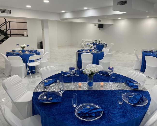 A spacious, well-lit event room set up with multiple round tables covered in blue tablecloths, arranged with white chairs and centerpieces.