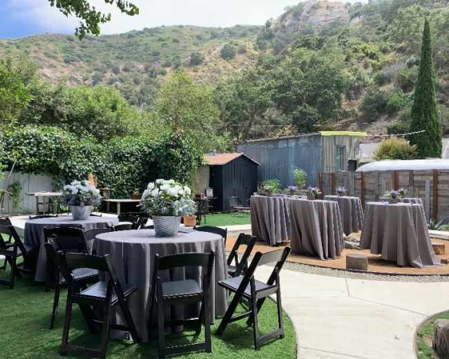 The scene shows an outdoor gathering area with several round tables covered in gray tablecloths, surrounded by greenery and a backdrop of hills.