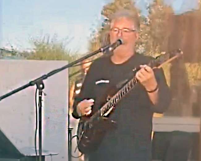 A musician plays an electric guitar and sings into a microphone while standing in front of a small audience.