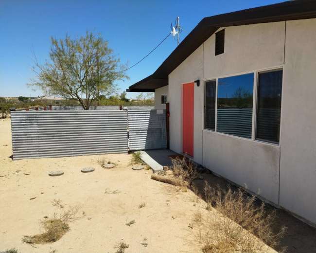 A modern house with a bright orange door is situated in a sandy landscape, surrounded by sparse vegetation and a corrugated metal fence.