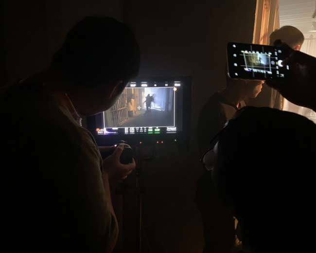 A group of people is observing a scene being filmed, with one person holding a camera and another using a phone to record the monitor displaying the footage.