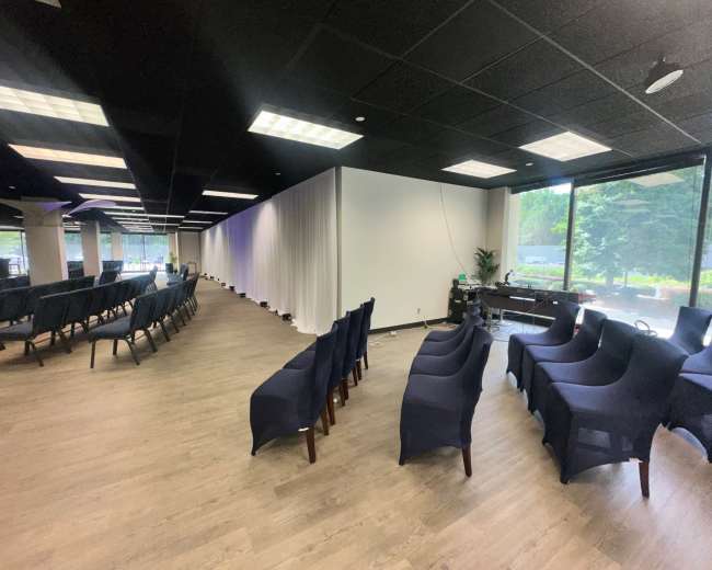 The image shows a spacious indoor area arranged with rows of navy blue chairs facing a white wall, accompanied by natural light from large windows.