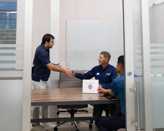 Two men are shaking hands in a glass-walled conference room labeled "ROVER ROOM," while a third man sits at the table with a box.