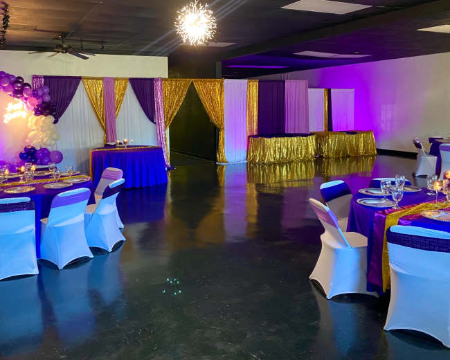 A decorated event space with purple and gold table settings, balloons, and a draped backdrop.