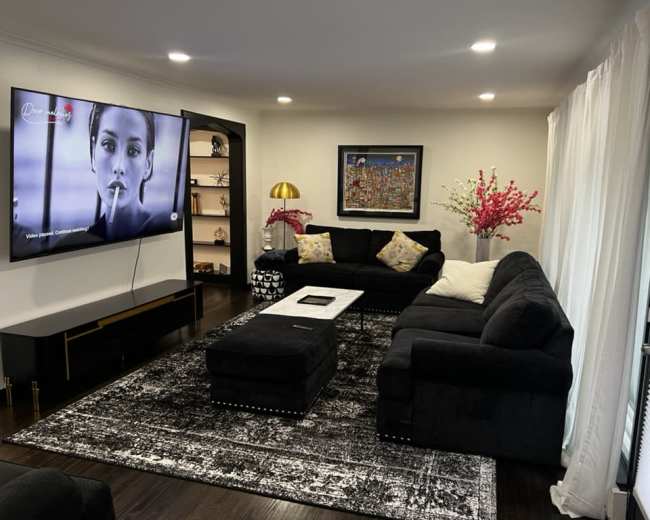 The living room features a black sectional sofa, a large television mounted on the wall, a coffee table in the center, and decorative elements including a gold lamp and a framed painting.