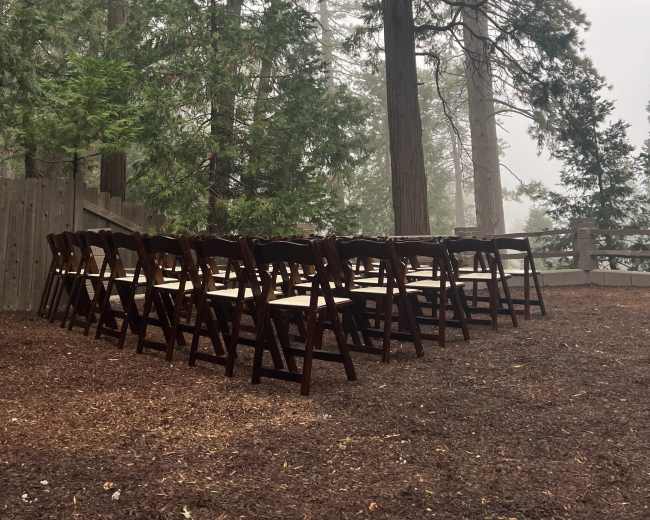 A row of wooden chairs is set up on a forested area surrounded by tall trees and hazy visibility.