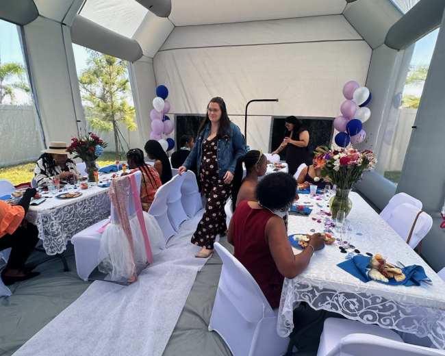 A woman in a denim jacket walks through a decorated tent where a small group of people are seated at tables, enjoying food and drinks.