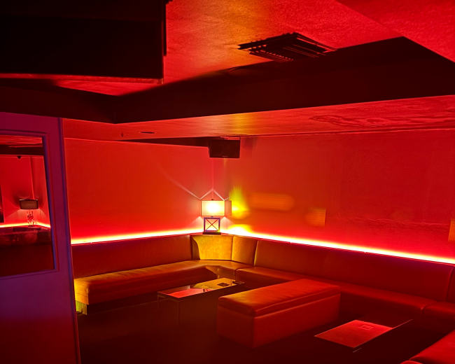 The image shows a dimly lit lounge with red lighting, featuring sofas arranged in a circular layout and low tables.