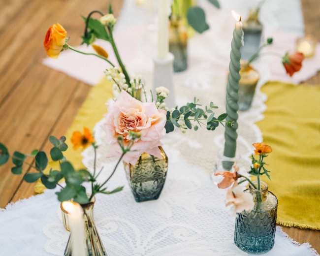 A long banquet table is adorned with floral arrangements, candles, and cloth runners in a variety of colors.