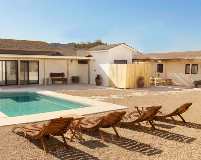 A modern white house with a pool and wooden lounge chairs is set in a gravel backyard surrounded by desert landscape.