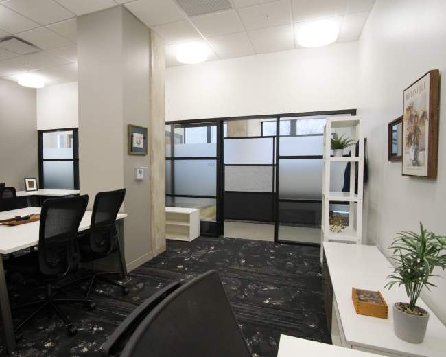 The image shows a modern office space featuring white desks, black office chairs, and a glass partition with a small shelf displaying plants and decor.