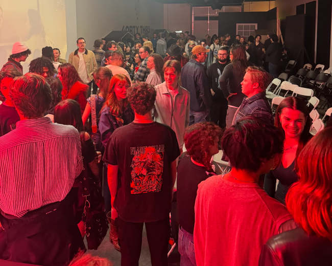 A crowded indoor space shows a diverse group of people gathered, with illuminated lanterns hanging from the ceiling.